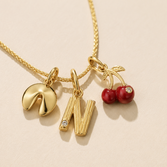 Gold necklace with three charms: a fortune cookie, letter 'N', and red cherries on a beige background.