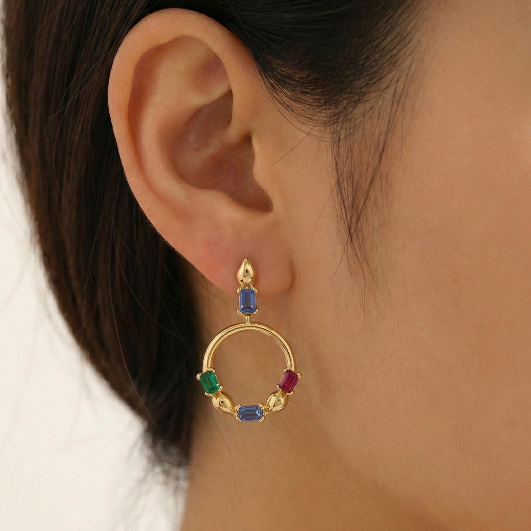 Gold hoop earring with colorful gemstones worn by a person.
