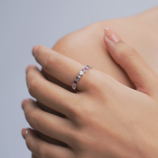 A delicate silver band ring adorned with small alternating pink and clear gemstones, shown worn on a person's finger with the other hand resting on the shoulder.