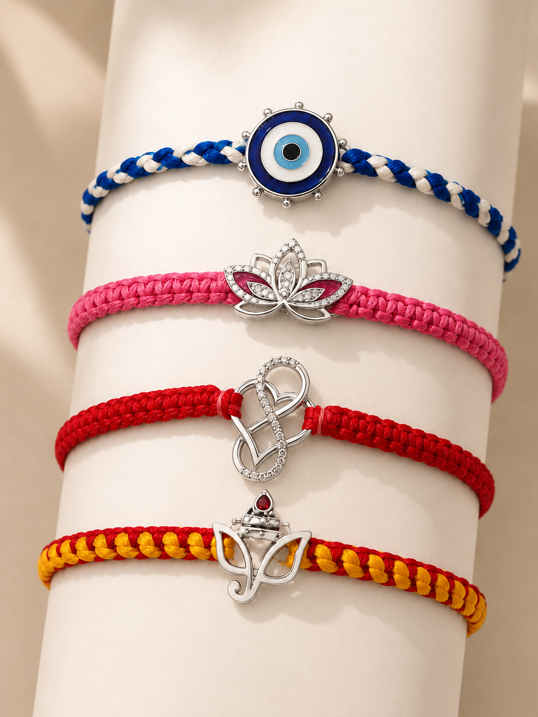 Four braided cord bracelets stacked on a white cylindrical display stand. From top to bottom: a blue and white bracelet with a circular evil eye charm, a pink bracelet with a silver lotus charm, a red bracelet with a silver infinity symbol, and a yellow and red bracelet with a silver crown charm.