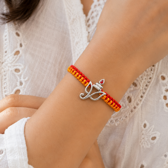 A close-up of a person's wrist wearing a handmade braided bracelet in red and orange with a silver-toned lotus charm.