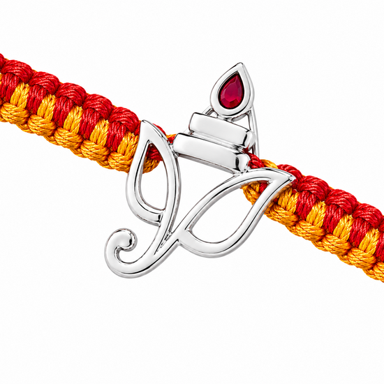 A close-up of a braided cord bracelet in red and yellow with a polished silver-toned diya pendant featuring a red teardrop stone, set against a plain white background.