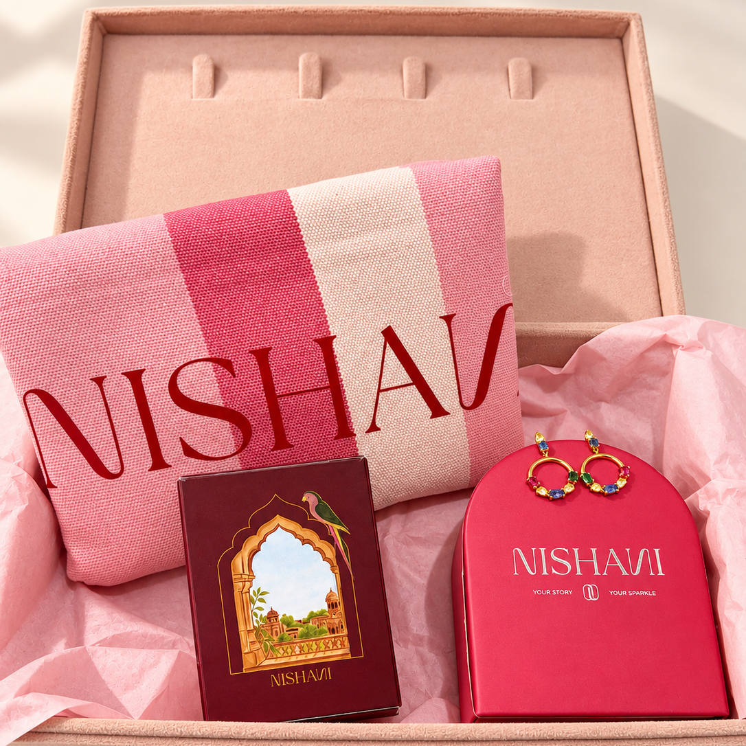 A NISHAVI gift set arranged in a blush pink presentation box lined with pink tissue paper. The set includes a pink and white striped scarf with red NISHAVI lettering, a dark red book or booklet with an arched window illustration, and a bright pink jewelry box with gold-tone earrings resting on top.