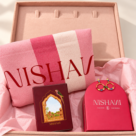 A NISHAVI gift set arranged in a blush pink presentation box lined with pink tissue paper. The set includes a pink and white striped scarf with red NISHAVI lettering, a dark red book or booklet with an arched window illustration, and a bright pink jewelry box with gold-tone earrings resting on top.