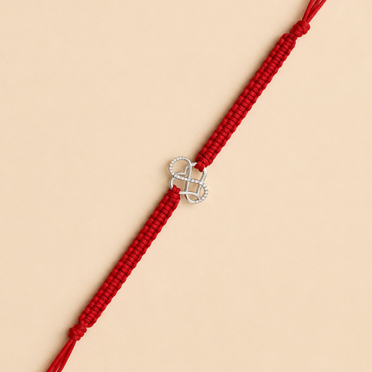 A red braided cord bracelet with a silver infinity symbol charm set with small clear stones, displayed diagonally on a solid beige background.