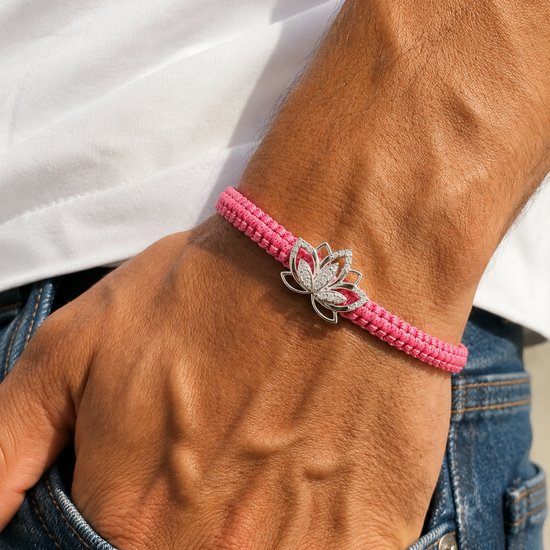 A close-up of a person's wrist wearing a bright pink beaded bracelet with a silver lotus flower charm, paired with a white shirt and blue jeans.