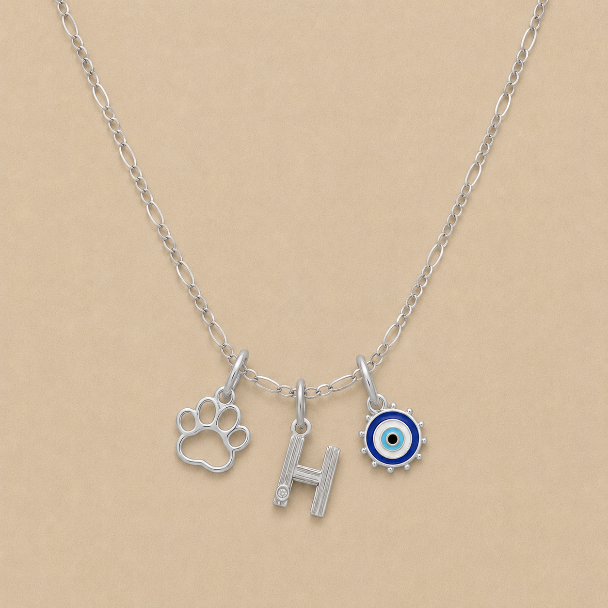 Silver necklace with paw print, letter 'H', and evil eye charm on a beige background